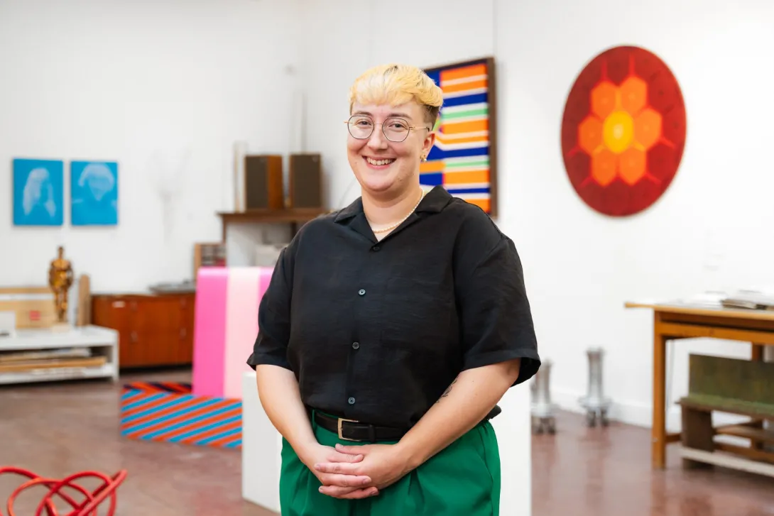 Ash Slemming is photographed standing and smiling while wearing a black-collared shirt and green pants. A number of artworks can be seen hanging on the wall in the background. Photo credit: Austin Kamenz.