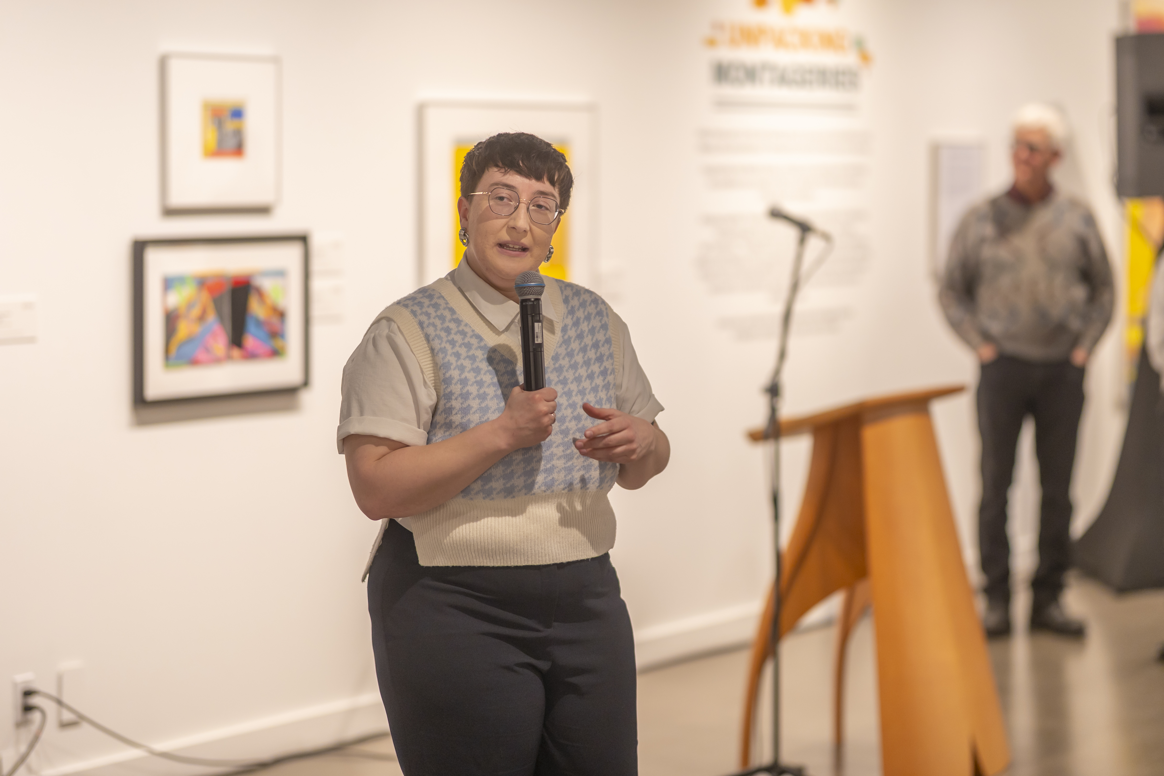 Ash Slemming speaks about the Montageries exhibition while holding a microphone and wearing a blue and white sweater vest.