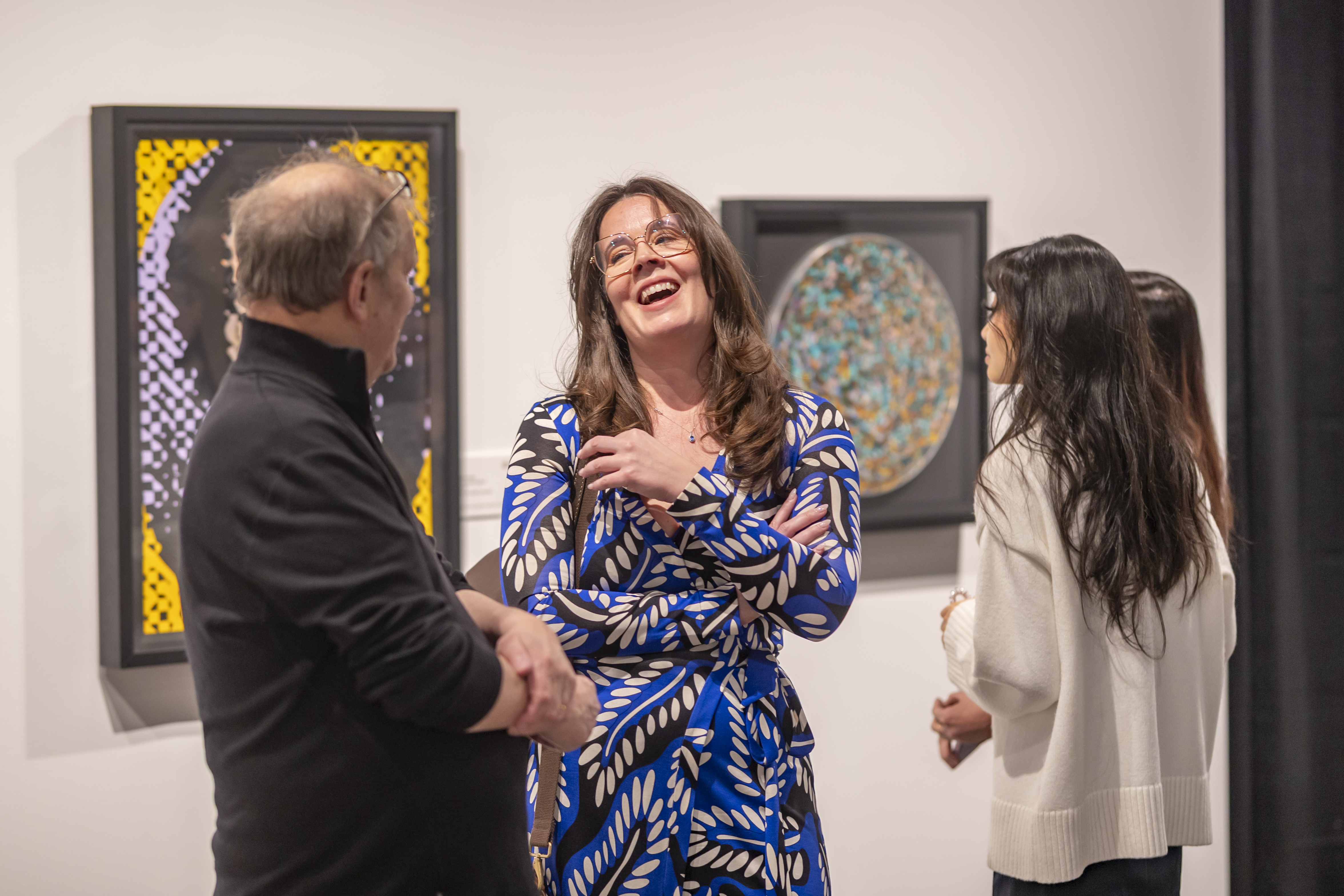 Three guests, one man and two women, share a moment. A brown-haried woman with glasses can be seen smiling while wearing a blue dress.