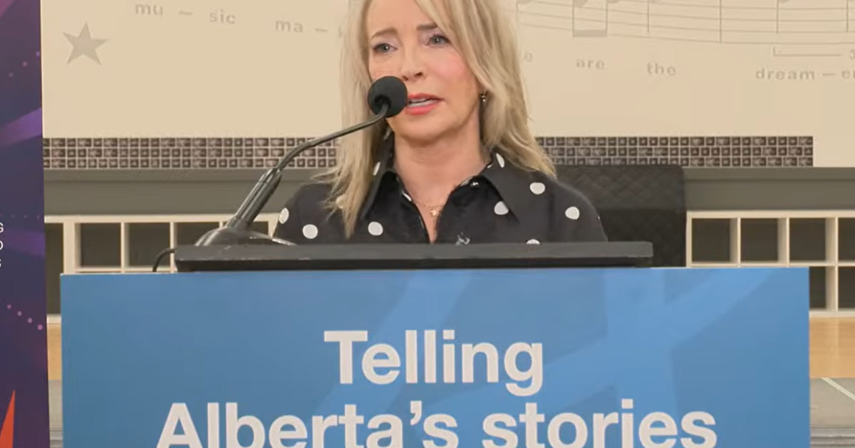 Cynthia P. Moore is standing behind a podium with a blue sign bearing the text "Telling Alberta's stories" and the Government of Alberta logo. Cynthia has blonde hair and is wearing a black blouse with white polka dots on it.