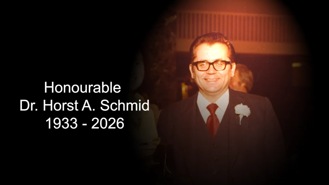 An archival photo of Horst Schmid, who is wearing glasses with a suit, red tie, and a corsage pinned to his suit jacket. Text accompanying the photo reads Honourable Dr. Horst A. Schmid 1933 – 2026.