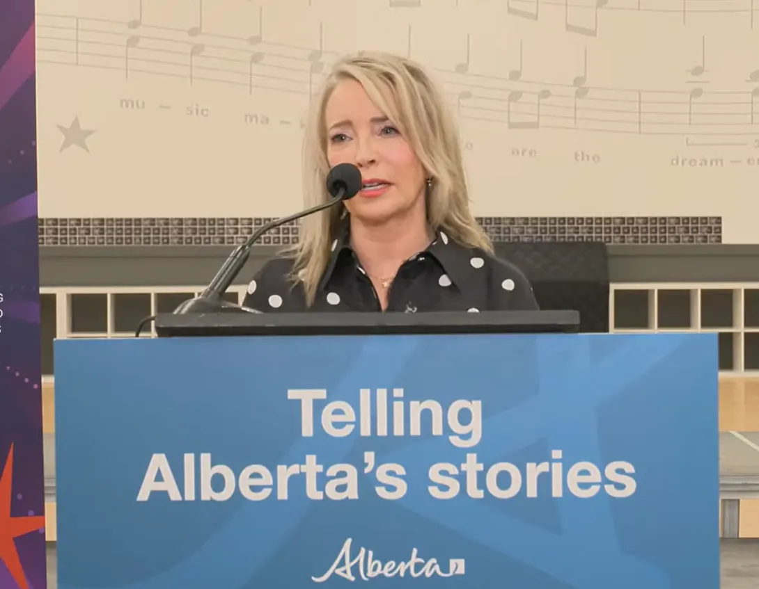 Cynthia P. Moore is standing behind a podium with a blue sign bearing the text "Telling Alberta's stories" and the Government of Alberta logo. Cynthia has blonde hair and is wearing a black blouse with white polka dots on it.