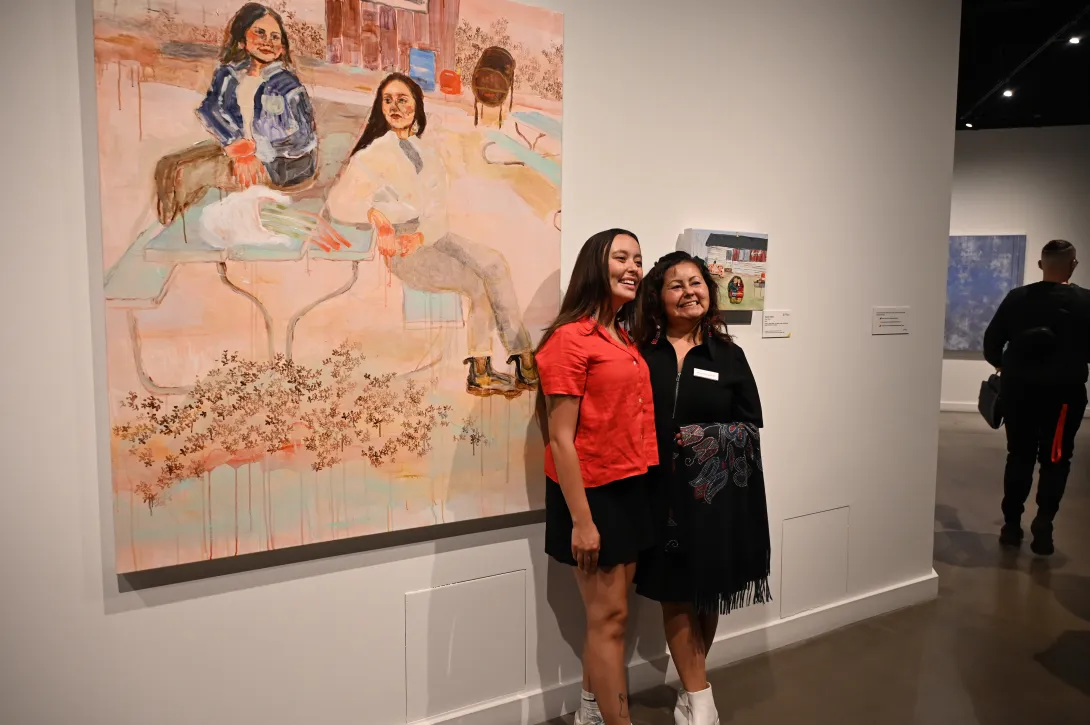 Kiona Callihoo Ligtvoet and Maureen Callihoo pose side by side beside their respective artworks. Kiona is wearing a red blouse and black skirt while Maureen is wearing a black jacket.